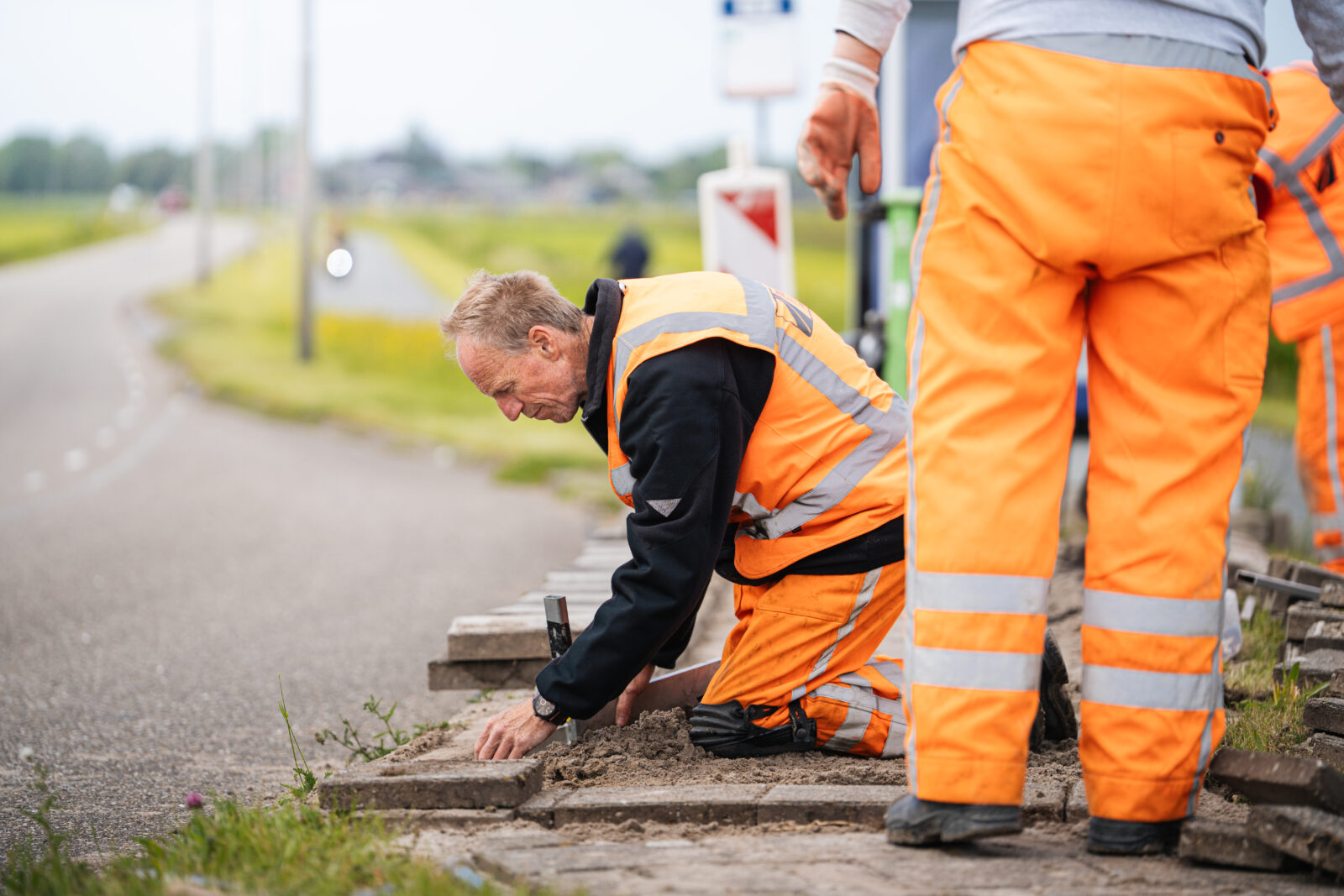 Medewerker van Ouwejan & F. de Bruijn voert bestratingswerkzaamheden uit langs een weg.