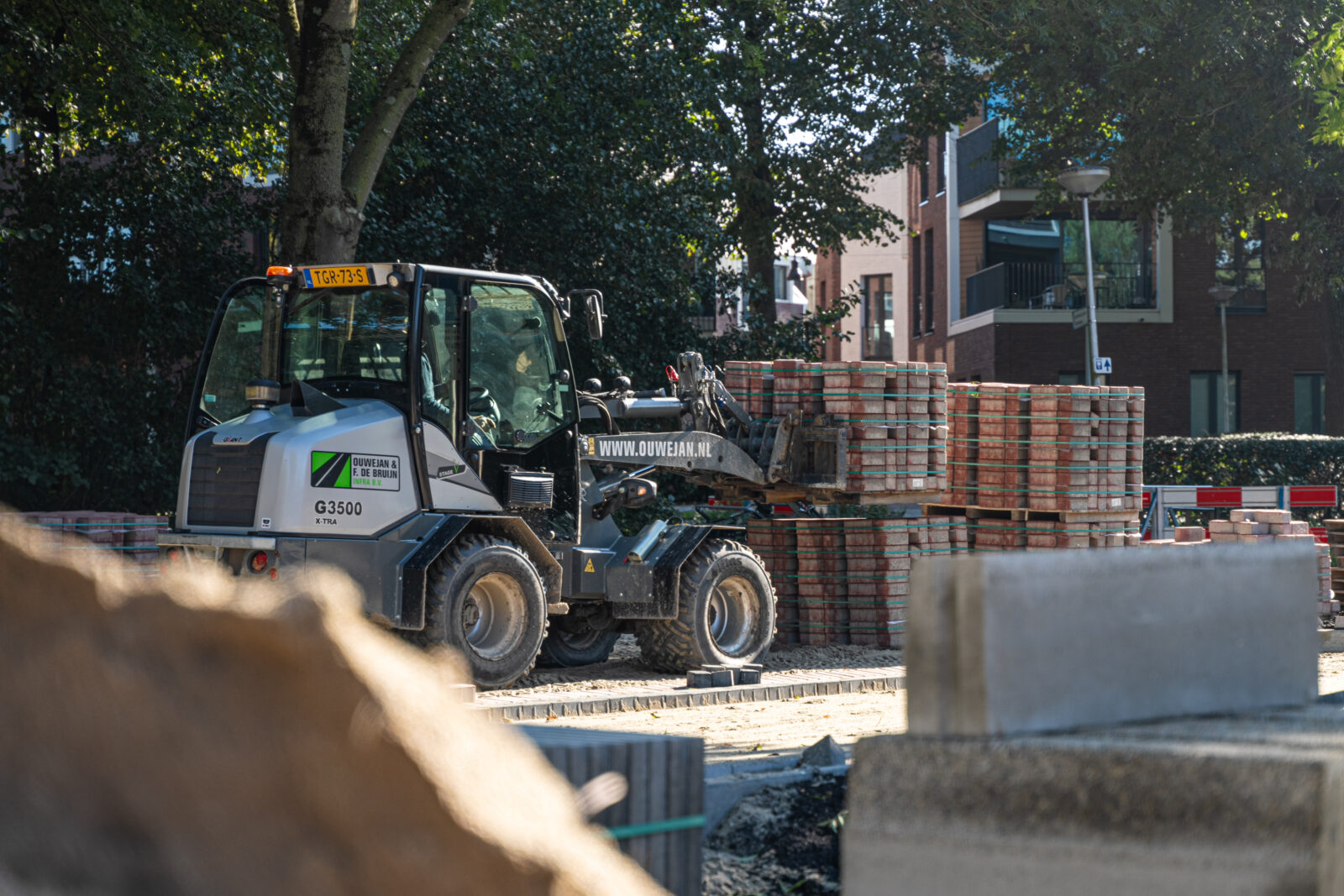 Medewerker van Ouwejan & F. de Bruijn transporteert pallets met bestratingsstenen op een projectlocatie.